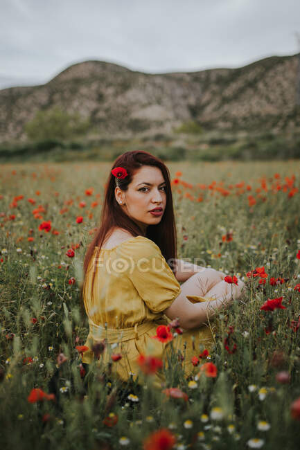 Pensive attraktive rothaarige erwachsene Dame in gelbem Kleid mit rotem Mohn im Haar und roten Lippen, die über die Schulter in die Kamera schauen, während sie allein auf einer verschwommenen grünen Wiese mit roten und weißen Blumen vor Hügeln unter grauem bewölkten Himmel sitzt — Stockfoto