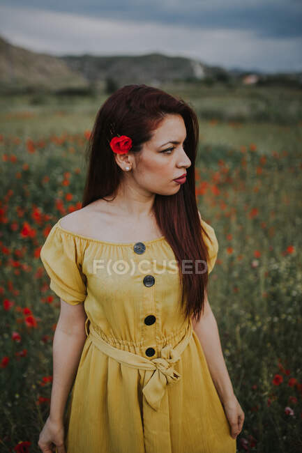 Attraktive rothaarige erwachsene Frau in gelbem Kleid mit rotem Mohn hinter dem Ohr schaut weg, während sie tagsüber allein auf einer verschwommenen grünen Wiese mit roten Blumen vor Hügeln unter grauem bewölkten Himmel steht. — Stockfoto
