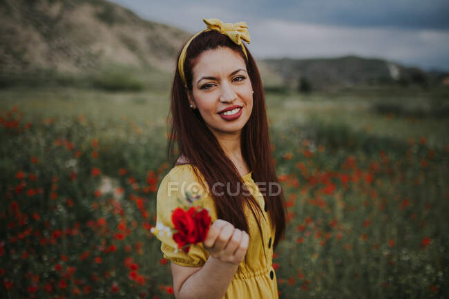Femme adulte aux cheveux rouges attrayants et souriants en robe jaune et bandeau avec lèvres rouges tenant bouquet avec rose rouge et regardant la caméra tout en restant seul dans un pré vert flou avec des fleurs rouges contre les collines sous un ciel nuageux gris pendant la journée — Photo de stock