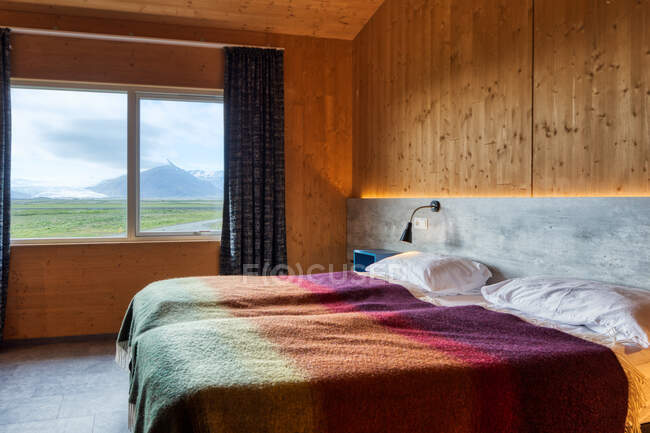 Две аккуратные удобные кровати с белыми подушками и теплыми красочными одеялами в светлой уютной комнате с желтыми стенами напротив окна с голубыми шторами с видом на сельскую местность — стоковое фото