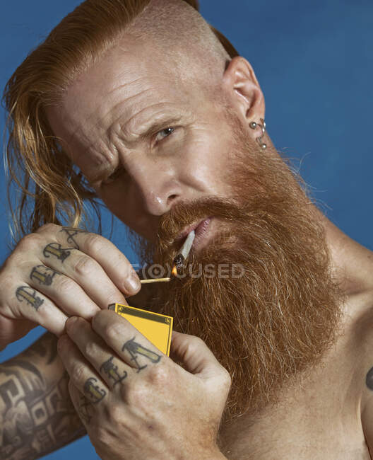 Продумана червоно-волохата бородата сорочка має татуювання на пальцях і руках, а з сережками у вухах чоловіка з сірими очима та підрізаною зачіскою, що тримає сірникову коробку та освітлювальну сигарету, підкидаючись та дивлячись на камеру на ізольованому синьому фоні — стокове фото