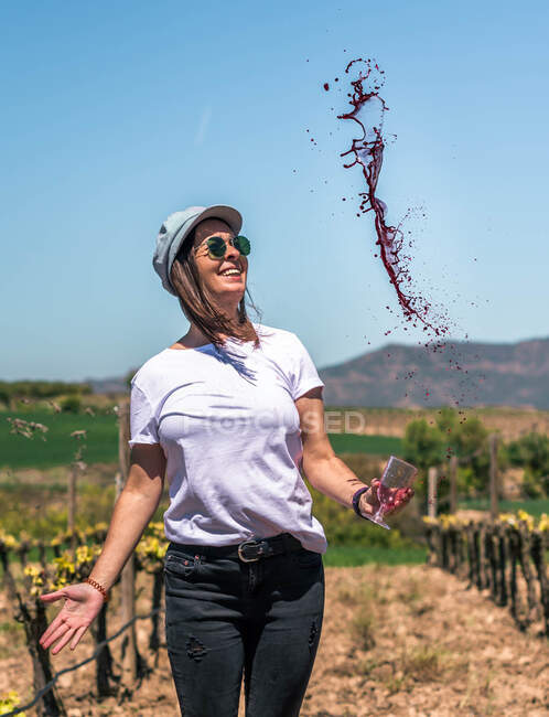 Turista piacevole allegro in berretto e occhiali da sole che ammirano gocce di vino rosso spruzzate su sfondo di campo verde — Foto stock