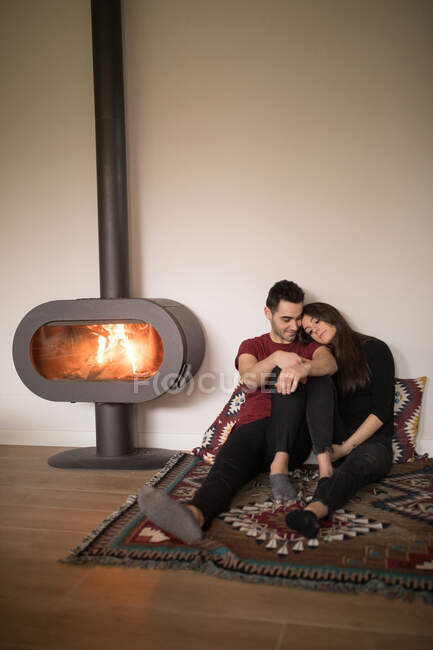 Pleased adult woman in casual clothes looking away and dreaming while sitting on floor and lying on shoulder of boyfriend in love near fireplace on colorful carpet against white wall at home — Stock Photo