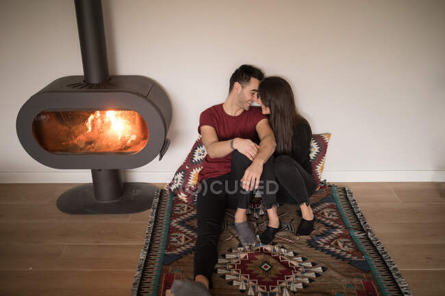Casal feliz em roupas casuais abraçando uns aos outros sentado no chão perto da lareira no tapete colorido contra a parede branca em casa — Fotografia de Stock