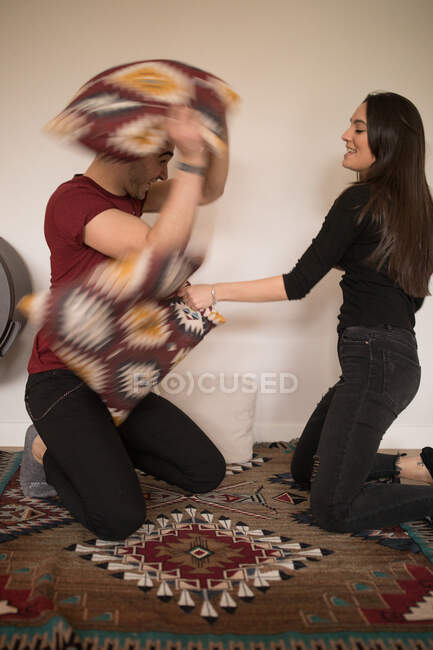 Side view of cheerful adult couple in casual clothes looking at each other and fighting pillows while standing on knees and playing on colorful carpet against white wall at home — Stock Photo