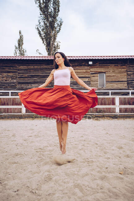 Художественная женщина в красной юбке прыгает в песчаном корпусе возле фермерского сарая при дневном свете — стоковое фото