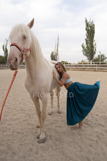 Seitenansicht einer anmutigen Frau in buntem langen Rock mit hochgeschlagenem Bein, die ein weißes Pferd umarmt, das noch am sandigen Gehege im Hippodrom steht — Stockfoto