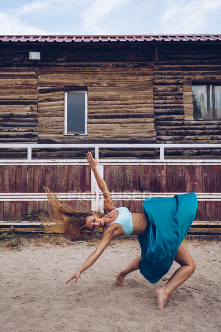 Vue latérale de la femme artistique en jupe longue ondulée dansant et équilibrant à l'enceinte sablonneuse par la grange de la ferme en plein jour — Photo de stock