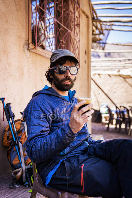 Homem relaxado em roupas quentes e óculos de sol bebendo bebida enquanto sentado na cadeira ao lado da mochila no terraço ensolarado da rua — Fotografia de Stock