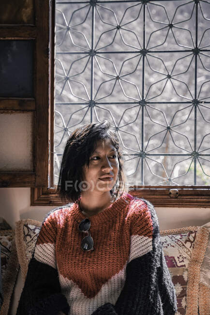 Нейтральная этническая женщина в теплом свитере смотрит, сидя на диване, украшенном окном при дневном свете — стоковое фото