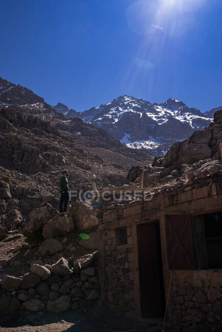 Viajante masculino olhando e em pé pela casa de pedra cercada por encostas rochosas no dia ensolarado — Fotografia de Stock