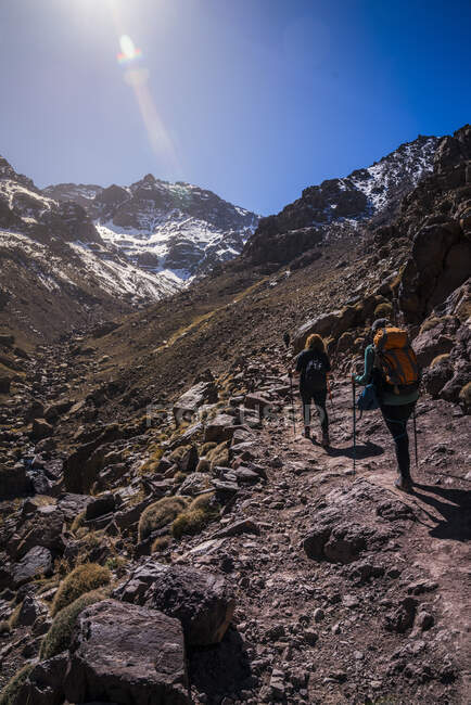 Rückansicht einer Gruppe von Menschen mit Rucksäcken, die mit Wanderstöcken auf felsigem Pfad am Gebirge entlang wandern — Stockfoto