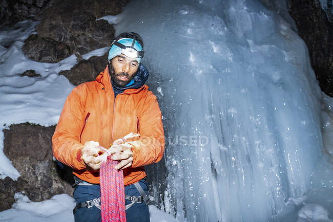 Bearded man in warm clothing with flashlight and equipment holding bundle of rope while standing by iced snowy cliff in darkness — Stock Photo