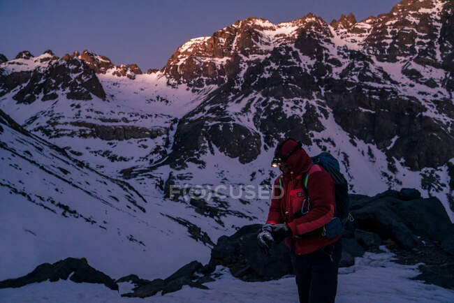 Людина в яскравій куртці з рюкзаком і ліхтариком відпочиває, стоячи в оточенні засніжених гірських вершин в сутінках — стокове фото