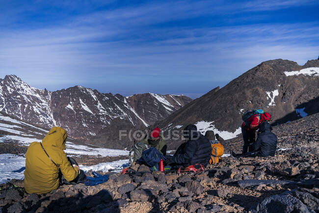 Back view group of people with backpacks in warm clothes sitting at rocky slope in sunny day — Stock Photo