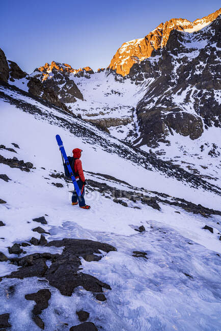Вид сбоку на человека в красочной куртке с рюкзаком и туристическим оборудованием, созерцающим, стоя на скалистом склоне при дневном свете — стоковое фото