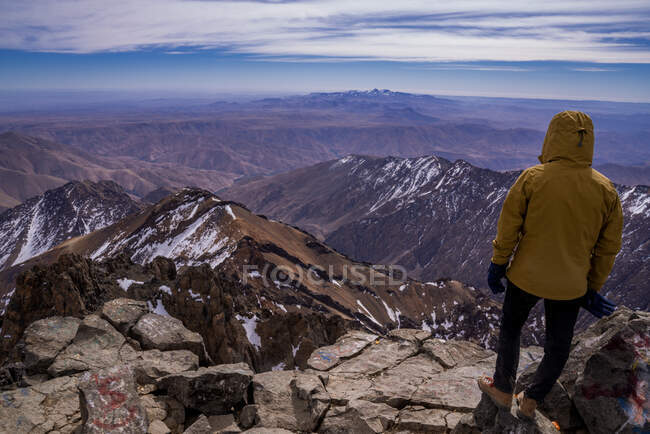 Rückansicht einer Person in warmer Jacke, die bei Tageslicht an einer Felswand steht und auf die Bergkette blickt — Stockfoto