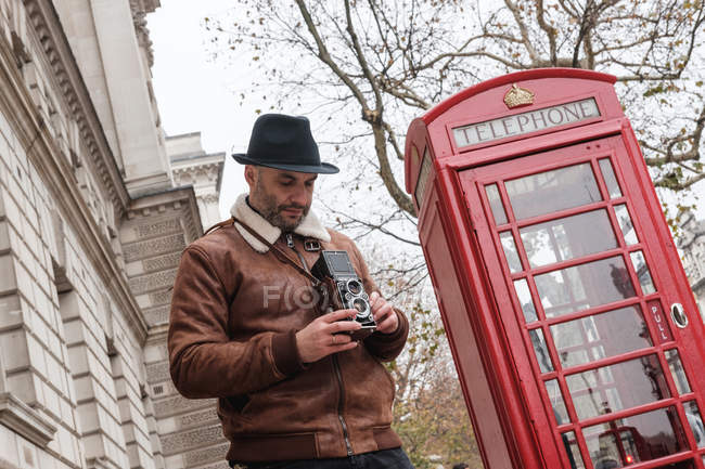 From below man with retro photo camera while standing on London street near traditional red telephone box — Stock Photo