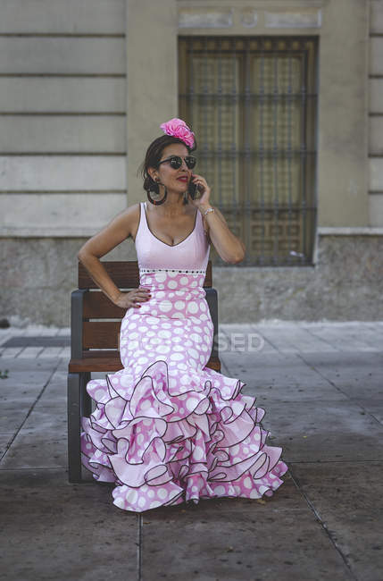 Ексцентрична жінка в барвисто-рожевому костюмі говорить на смартфоні, сидячи на пляжі вздовж літньої вулиці — стокове фото