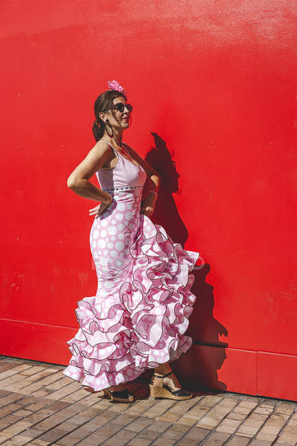 Vue latérale de femme excentrique gaie en costume rose coloré souriant et dansant par fond de mur rouge sur une journée ensoleillée — Photo de stock