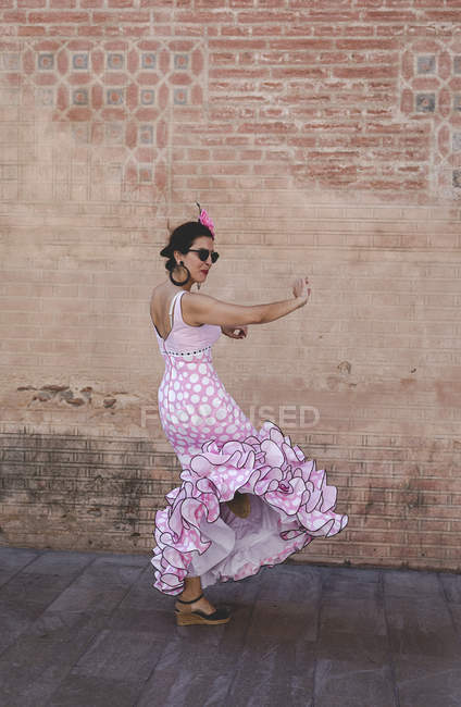 Боковой вид эксцентричной веселой женщины в красочном розовом костюме, улыбающейся и танцующей у кирпичной стены в солнечный день — стоковое фото