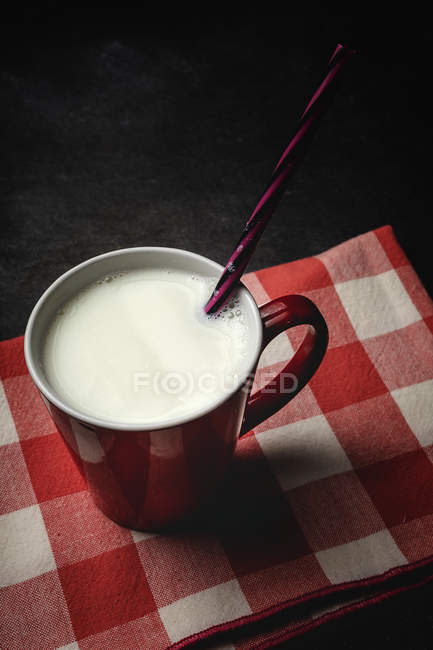 Coppa di latte bianco con brillante cannuccia a righe sul tavolo sopra sfondo nero e asciugamano a scacchi — Foto stock