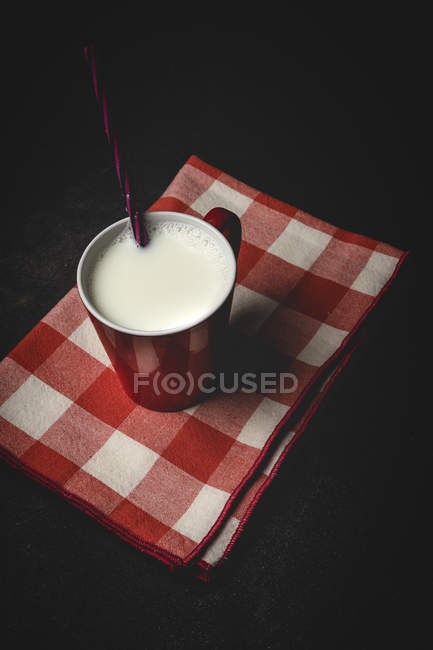Coppa di latte bianco con brillante cannuccia a righe sul tavolo sopra sfondo nero e asciugamano a scacchi — Foto stock