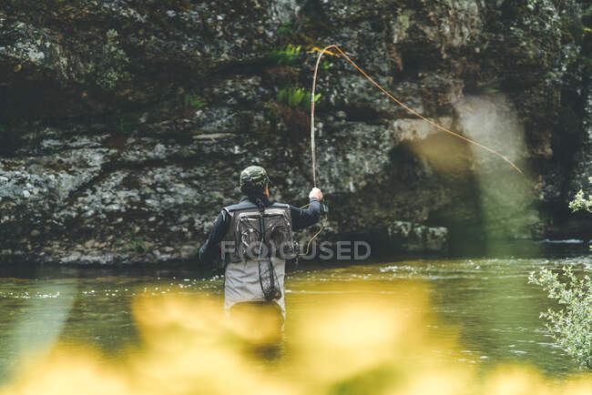 Back view of equipped man harling fish while standing in waders in mountain torrent by cliff and forest — Stock Photo