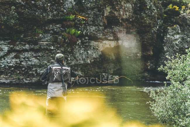 Vista trasera del hombre equipado harling pescado mientras está de pie en vadeadores en torrente de montaña por acantilado y bosque - foto de stock