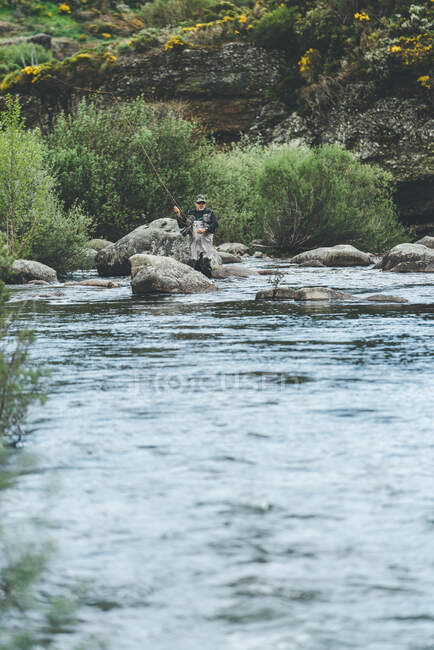 Обладнана людина, що ловить рибу, стоячи всередині води в річковому потоці в гірському потоці за скелею та лісом — стокове фото