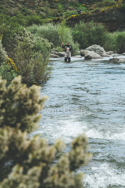 Equipped man harling fish while standing inside water in river stream in mountain torrent by cliff and forest — Stock Photo