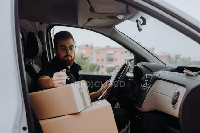 Correio em óculos preparando pacotes para transporte enquanto sentado e marcando caixas no carro em fundo turvo durante o dia — Fotografia de Stock