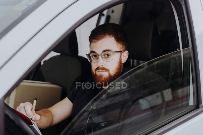 Homme se concentrant et vérifiant les documents tout en étant assis derrière le volant dans la cabine de voiture pendant la journée sur fond flou — Photo de stock