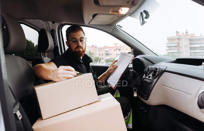 Kurier in Brille bereitet Pakete für den Transport vor, während er tagsüber im Auto sitzt und Kisten markiert — Stockfoto