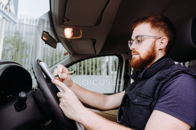 Homem focando e verificando documentos enquanto sentado ao volante na cabine do carro durante o dia no fundo turvo — Fotografia de Stock