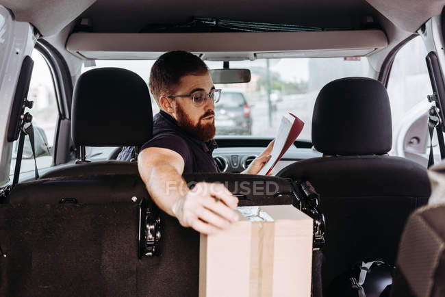 Correio em óculos preparando pacotes para transporte enquanto sentado e marcando caixas no carro em fundo turvo durante o dia — Fotografia de Stock