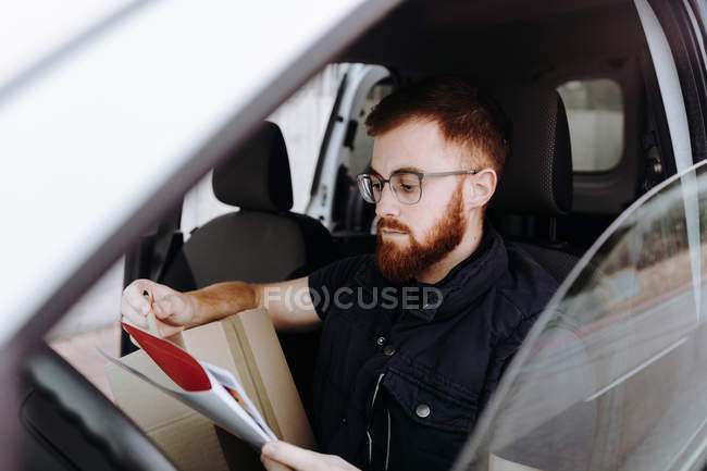 Чоловік фокусує і перевіряє документи, сидячи за кермом в салоні автомобіля вдень на розмитому тлі — стокове фото