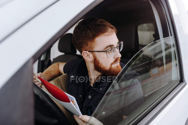 Чоловік фокусує і перевіряє документи, сидячи за кермом в салоні автомобіля вдень на розмитому тлі — стокове фото