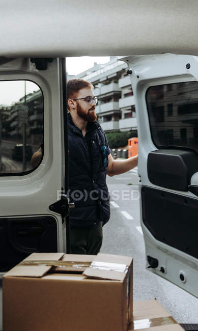Courier in glasses carefully unloading cardboard boxes from car for further delivery to customer on blurred background during daytime — Stock Photo