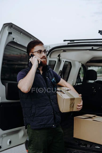 Курьер в очках держит коробки и делает предупреждение клиенту для дальнейшей доставки, стоя возле багажника автомобиля и глядя в сторону в дневное время — стоковое фото