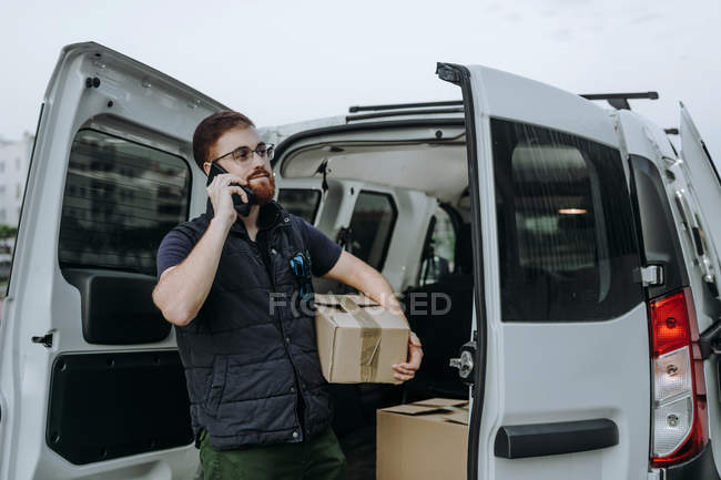 Courier in glasses holding boxes and making warning call to customer for further delivery while standing near car trunk and looking away during daytime — Stock Photo
