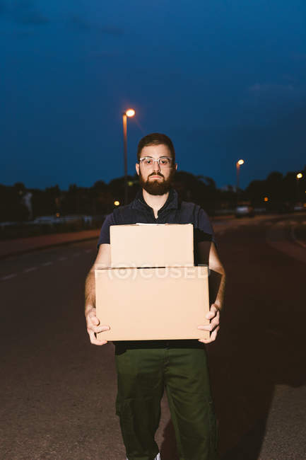 Uomo in occhiali che trasportano scatole mentre in piedi sulla strada e guardando la fotocamera vicino lampione incandescente in serata su sfondo sfocato — Foto stock