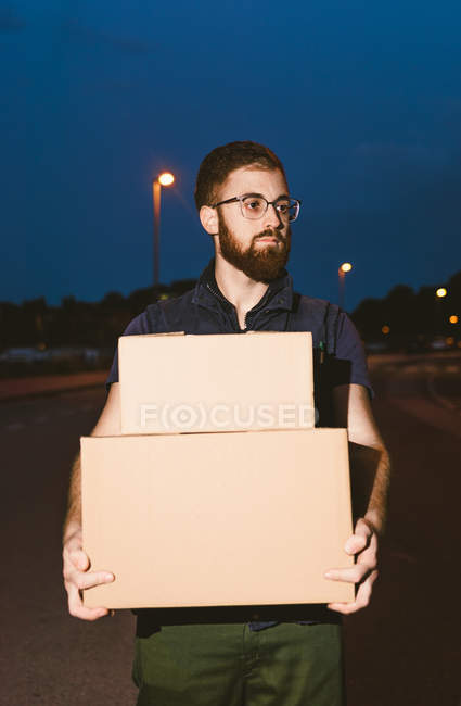 Mann mit Brille trägt Kisten, während er auf der Straße steht und abends in der Nähe glühender Straßenlaternen auf verschwommenem Hintergrund wegsieht — Stockfoto