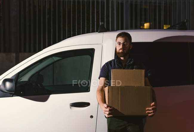 Uomo in occhiali pensando e guardando lontano mentre in piedi vicino alla macchina e tenendo scatole di cartone in serata — Foto stock