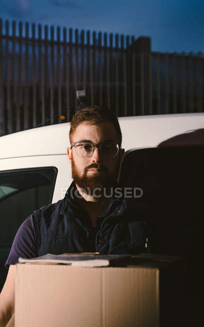 Hombre barbudo adulto en gafas pensando y mirando la cámara mientras está de pie cerca del coche y sosteniendo cajas de cartón por la noche - foto de stock