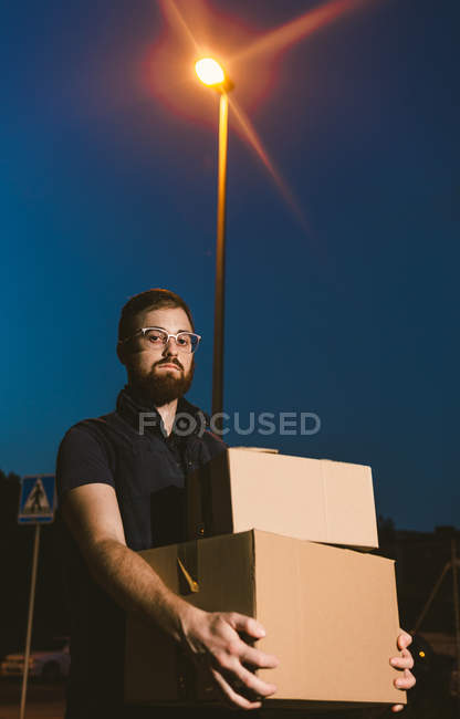 Человек в очках, несущий коробки, стоя на улице и глядя на камеру возле светящегося уличного фонаря вечером на размытом фоне — стоковое фото