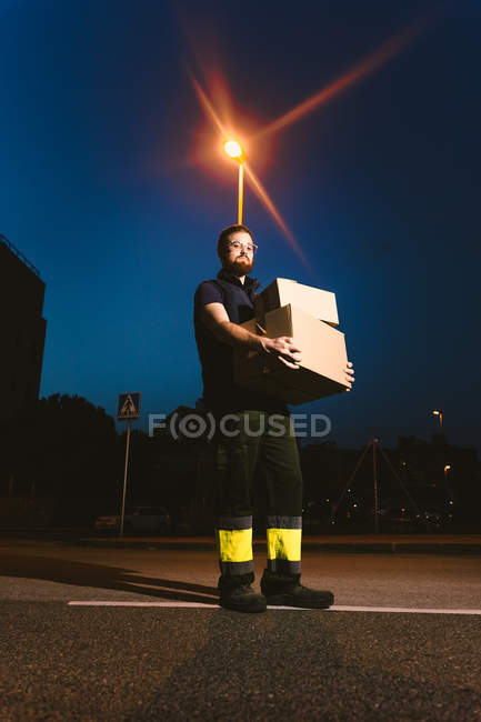 Человек в очках, несущий коробки, стоя на улице и глядя на камеру возле светящегося уличного фонаря вечером на размытом фоне — стоковое фото