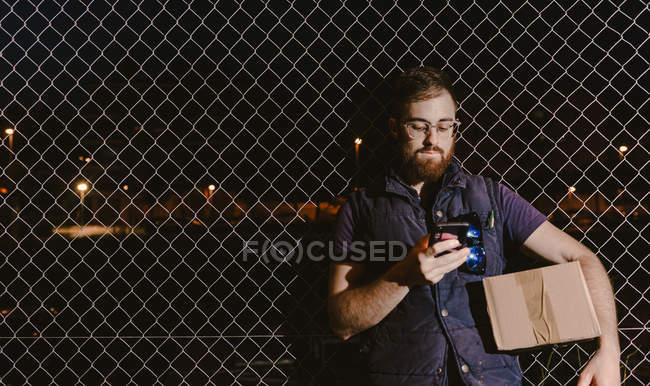 Courrier dans des lunettes avec des boîtes regardant la caméra tout en se tenant debout et appuyé sur une clôture en maille parler sur le téléphone mobile dans la rue la nuit — Photo de stock