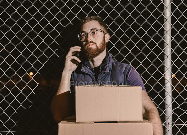 Курьер в очках с коробками, смотрящими в камеру, стоя и опираясь на сетку забора, разговаривая по мобильному телефону на улице ночью — стоковое фото