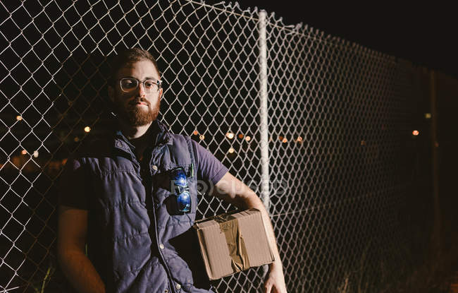 Courier in glasses with boxes looking at camera while standing and leaning on mesh fence on street at night — Stock Photo